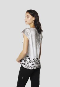 Īsām piedurknēm krekls baltā un pelēkā gradientā ar melnu ziedu apdruku apakšmalā. Gluds audums ar maigu tekstūru.