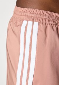 Lyserøde træningsshorts lavet af glat stof, med en elastisk talje og tre hvide lodrette striber langs siden.