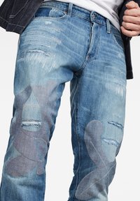 Jeans de mezclilla azul desgastados que presentan desgaste visible, recortes parcheados y acentos bordados en los paneles frontales para un detalle adicional.