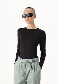 Top preto de mangas longas combinado com calças verdes claras de cintura alta, apresentando um laço na cintura e bolsos laterais. Óculos de sol escuros completam o visual.