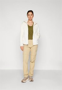 Baltas flisinis striukės užtrauktukas ant alyvuogių žaliumo maikutės, derinamas su smėlio spalvos krovinių kelnėmis ir šviesiai rudu sportbačiais, turinčiais reguliuojamas rankogalius ir kišenes.