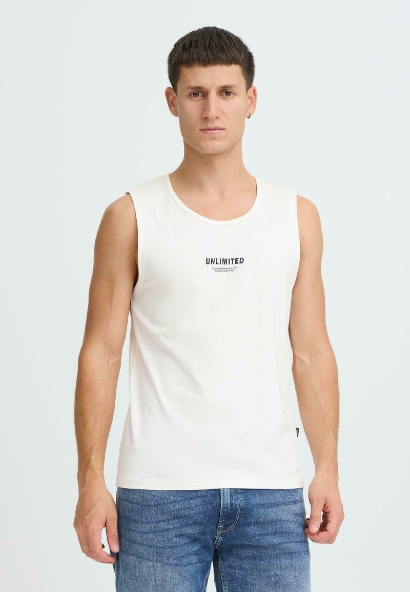 T-shirt blanc sans manches avec un col rond, arborant une inscription noire "UNLIMITED" sur la poitrine. Porté avec un jean en denim bleu.