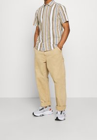 Korte mouw, gekleed shirt met verticale beige en witte strepen. Gecombineerd met tan cargobroek en witte sneakers met zwarte en oranje accenten.