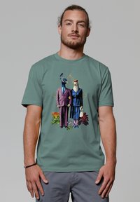 Groen T-shirt met korte mouwen, met een kleurrafisch van twee vogels in formele kleding, tegen een bloemig achtergrond met geometrische accenten.