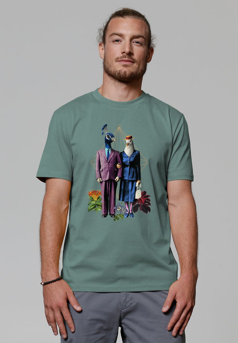 Groen T-shirt met korte mouwen, met een kleurrafisch van twee vogels in formele kleding, tegen een bloemig achtergrond met geometrische accenten.