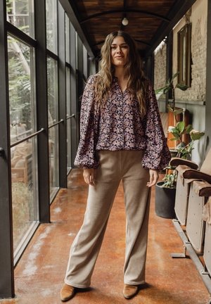 Paarse en roze gebloemde blouse met pofmouwen, gecombineerd met beige wijde broek en lichtbruine schoenen, gedragen door een model.