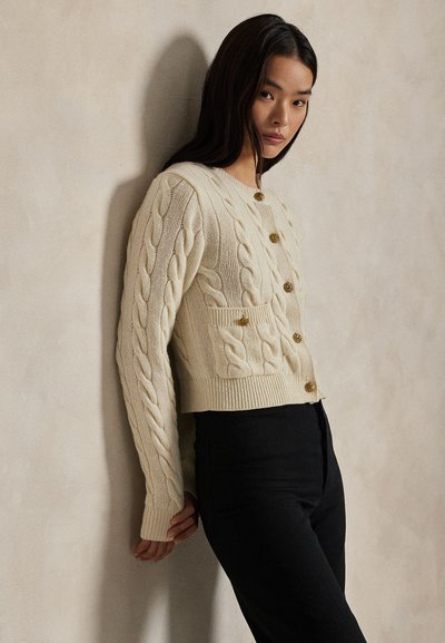 Vrouw leunend tegen een beige muur, draagt een crème kabelgebreid vest met gouden knopen en een zwarte broek, kijkt naar de camera.