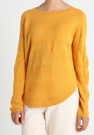 Langarmige, gestructureerde mosterdgele trui met een ronde hals, gedragen door een persoon in lichtgekleurde broek tegen een effen achtergrond.