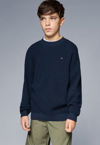 Granatowy sweter z dzianiny o fakturowanym wzorze, okrągły dekolt i ściągacze na mankietach. Mały detal logo na klatce piersiowej. Noszony z jasnymi spodniami.