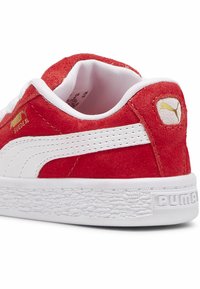Adidași roșii din suede cu accente albe, având o talpă texturată albă și un detaliu cu logo de aur pe călcâi. Design clasic de tip low-top.