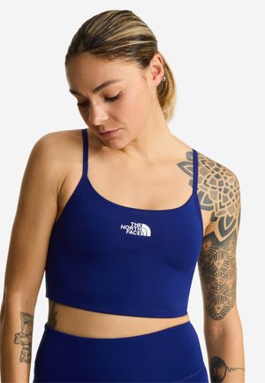 Mujer con tatuajes en el brazo izquierdo que lleva una camiseta corta azul con el logo de The North Face, mirando hacia abajo sobre un fondo blanco.