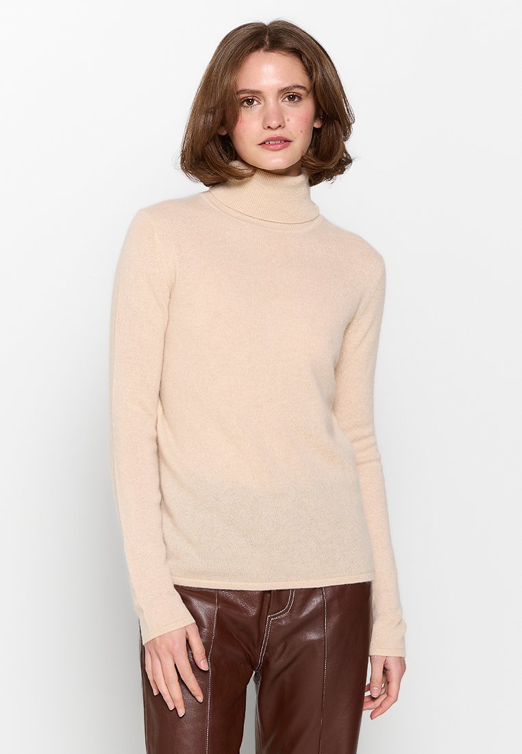 Beige Rollkragenpullover aus weichem Stoff, taillierte Passform, lange Ärmel, gerippter Kragen, kombiniert mit braunen Lederhosen, glatte Textur.