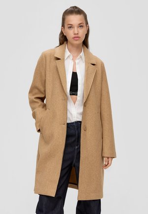 Camel-farbenem Wollmischmantel mit Reverskragen, zwei Knöpfen und Seitentaschen, getragen über einem weißen Hemd und einem gemusterten Top.