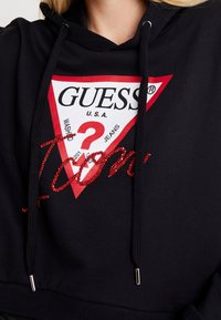 Sudadera con capucha negra que presenta un logotipo triangular en rojo y blanco con "GUESS" y un signo de interrogación. "I Am" está intrincadamente bordado en rojo.