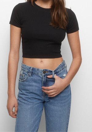 Femme portant un t-shirt noir coupé court et un jean bleu taille haute, tenant la ceinture d'une main, sur un fond uni.
