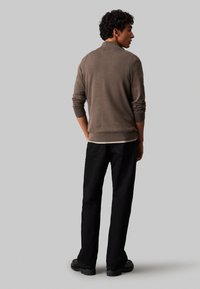 Brauner Strickpullover mit langen Ärmeln, gerippten Bündchen und einem runden Kragen, kombiniert mit schwarzen, geraden Hosen und schwarzen Schuhen.