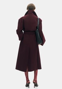 Bordeaux wollen trenchcoat met een ceintuur in de taille, brede mouwen en een hoge kraag. Gekozen met doorzichtige, gedessineerde rode panty's en zwarte hakken.