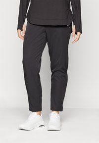 A person wearing a long-sleeve black top and black pants with white athletic shoes stands in a neutral setting, focused on their lower body.