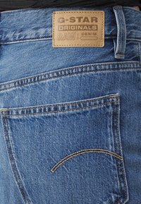 Svijetloplave traperice sa vidljivom stražnjom džepom. Imaju smeđi kožni etiketu s natpisom "G-STAR ORIGINALS RAW DENIM." Istaknuti detalji šivanja.