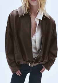 Giacca di pelle marrone con zip frontale, ampio colletto e maniche con polsini. Abbinata a una camicia bianca con bottoni e jeans scuri.
