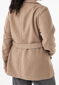 Person med lockigt hår iförd en beige ullkappa med bakrem och grå rutiga byxor, visad bakifrån.