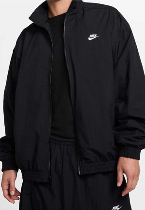 Man die een zwarte Nike-jas met rits draagt en bijpassende zwarte Nike-shorts met elastische tailleband, staand tegen een witte achtergrond.