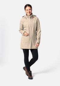 Beige waterdichte jas met een capuchon, rits aan de voorkant, twee zijzakken en verstelbare trekkoorden. Zwarte leggings en bruine wandelschoenen.
