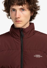 Burgundy vadderad jacka med hög krage, med dragkedja framtill och vit logotext på vänster sida. Slät tygstruktur.