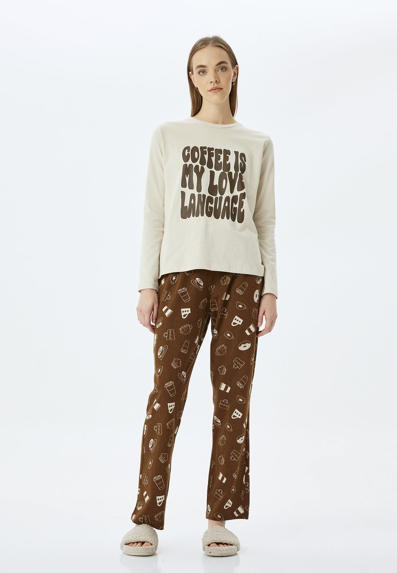 Camisa de manga larga color crema con texto en negrita, combinada con pantalones de pijama marrones que tienen un patrón de comida y bebida. El modelo lleva zapatillas beige.