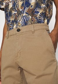 Beiga bomullbyxor med knapp och dragkedja, kombinerat med en kortärmad skjorta med ett blått, brunt och vitt bladmönster.