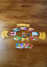 Układ planszy do gry z kolorowymi kartami z postaciami zwierząt, o okrągłym kształcie, teksturowanymi powierzchniami oraz elementami gry, w tym kostką i kolorowymi żetonami.