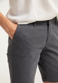 Una persona che indossa pantaloni corti grigi scuri e una camicetta bianca a trama, con una mano casualmente in tasca.
