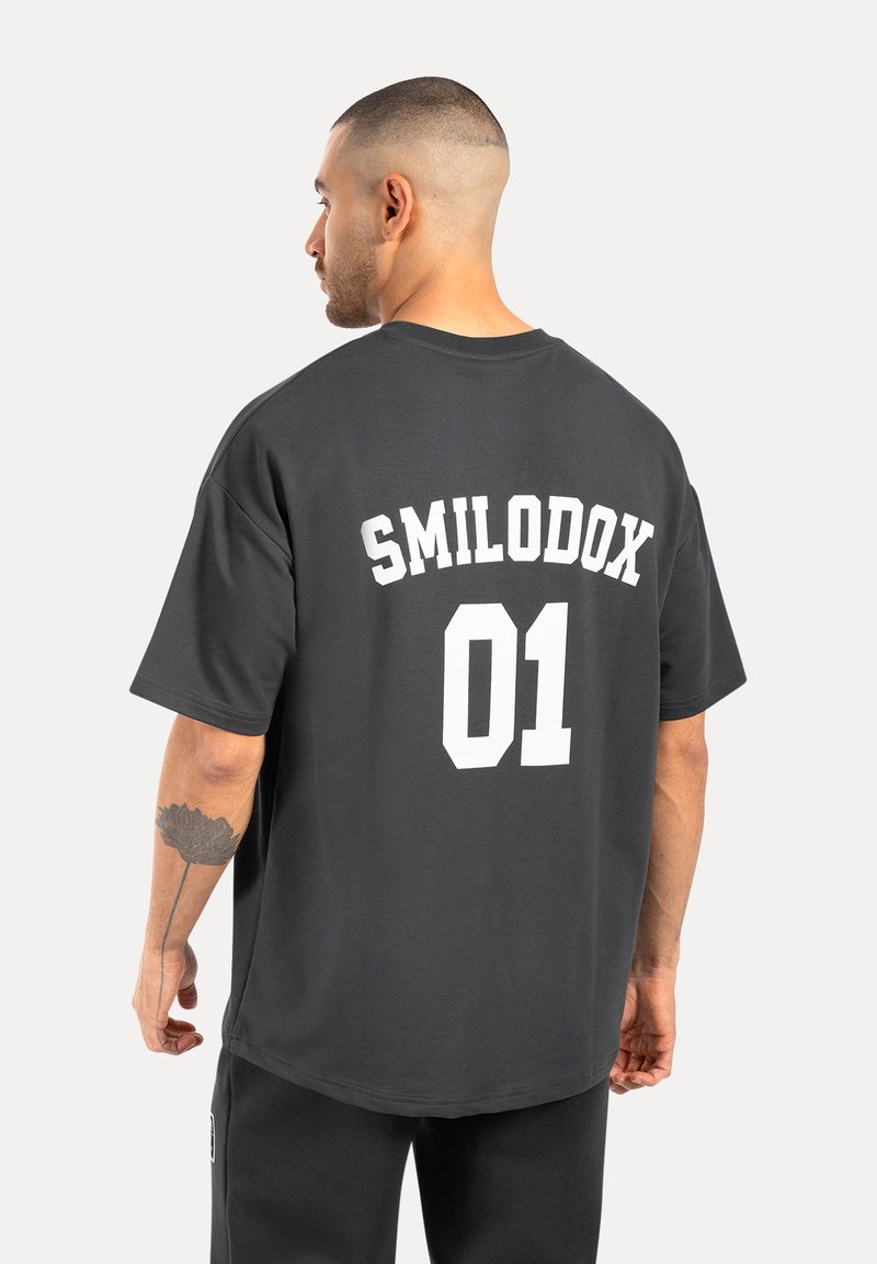 Grå oversized t-shirt med "SMILODOX" og "01" i stor hvid skrift på ryggen. Lavet af bomuld, med korte ærmer og rund halsudskæring.