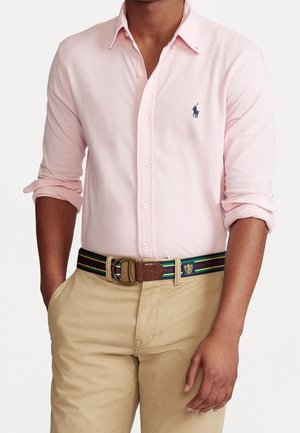 Mann trägt hellrosa Hemd mit Knöpfen, hochgekrempelte Ärmel, beige Hose und gestreiften Stoffgürtel mit Lederdetails.