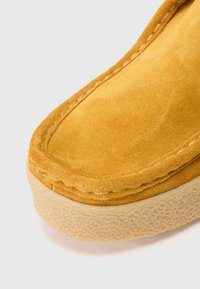 Sárga suede slip-on cipő, lekerekített orral, kontrasztos varrással a szélen, és textúrázott gumitalppal.