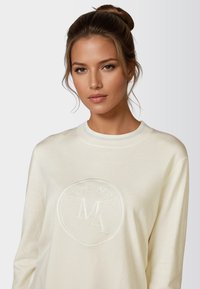 Cream-colored sweatshirt with ribbed neckline. Features an embossed circular logo in the center. Soft, textured fabric. Long sleeves.