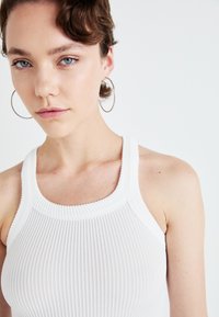 Débardeur blanc côtelé avec un col rond. Le tissu présente un motif texturé. Le mannequin porte des boucles d'oreilles en argent en forme de créole.