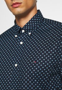 Camicia blu navy con bottoni, realizzata in cotone con un piccolo motivo floreale blu, caratterizzata da bottoni bianchi e un logo sul petto.