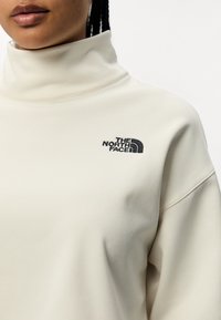 Kermanvärinen, korkeakauluksinen collegepaita, jossa on resorikuvio ja vasemmassa rinnassa musta kirjailtu logo. Rentoon tyyliin suunniteltu, väljä istuvuus.