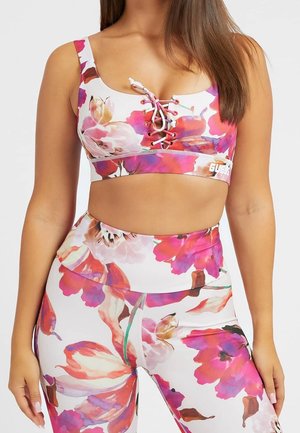 Vrouw in een bloemen sport-bh met een voorzijde met vetersluiting en bijpassende high-waist legging in roze, paars en witte tinten.