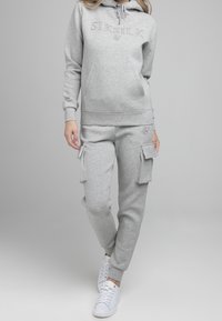 Mujer vistiendo una sudadera SikSilk gris clara y pantalones de chándal cargo con zapatillas blancas, de pie contra un fondo gris liso.