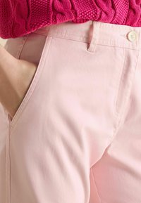 Gaiši rozā kokvilnas bikses ar gludu tekstūru, priekšējām kabatām un mazu baltu pogu aizdari jostasvietā.