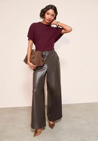 Burgundská oversized pletená blúzka v kombinácii s vysokými hnedými nohavicami z imitácie kože s širokými nohavicami a hnedými špičatými podpätkami. V ruke má hnedú kabelku.