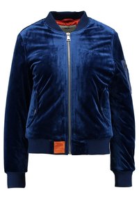 Veste bomber en velours bleu marine avec fermeture éclair au devant, poignets et ourlet côtelés, poches latérales, et une étiquette orange sur la ceinture.