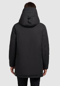 Osoba v černé vycpané bundě s vysokým límcem a kapucí, zobrazená zezadu na neutrálním světlém pozadí.