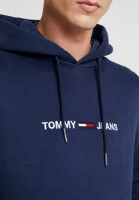 Närbild av en person som bär en marinblå hoodie med "TOMMY JEANS" broderat i vitt och en röd, vit och svart logotyp mellan orden.