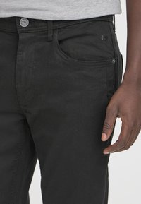 Črne denim hlače z gladko teksturo, ki imajo zaponko in sprednje žepke. Majhna oznaka je vidna blizu žepa.