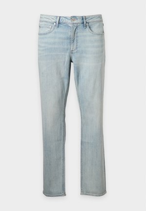 Mand iført en beige kortærmet strikket poloshirt, lyseblå jeans og sorte læderloafer står med den ene hånd i lommen.