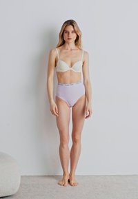 Beige bra with thin straps and high-waisted lavender underwear with a Calvins logo. Model has natural hair and stands on a light carpet.