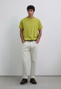 Homme debout avec les mains dans les poches, portant un t-shirt vert lime, un pantalon blanc cassé, une ceinture noire et des chaussures brun foncé devant un mur uni.